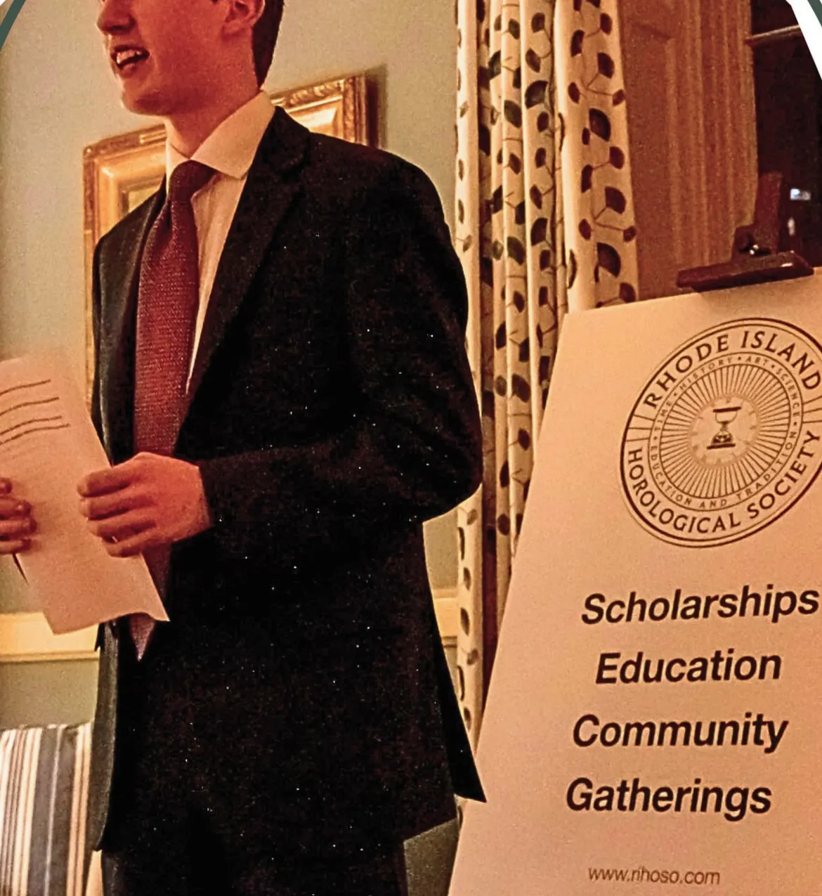 A young speaker addresses guests beside a RIHOSO banner reading Scholarships, Education, Community, Gatherings