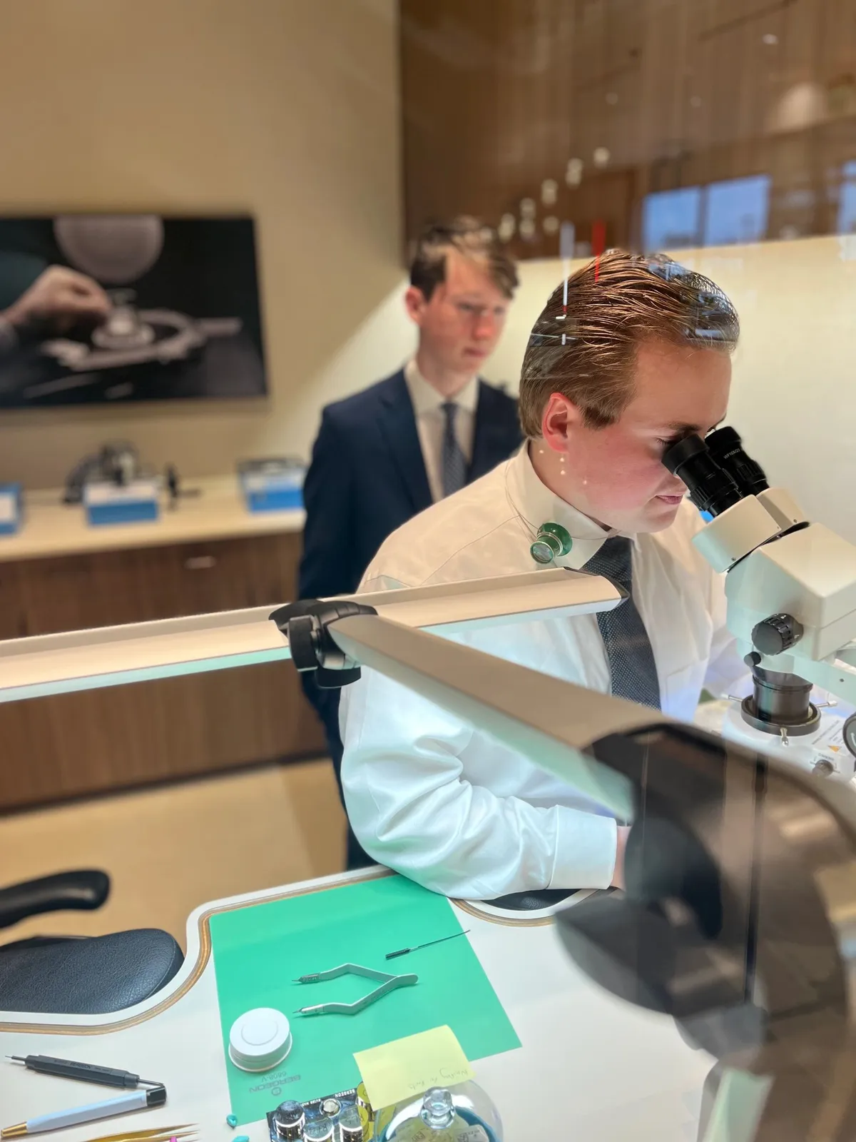 A watchmaker works at a microscope while a member looks on